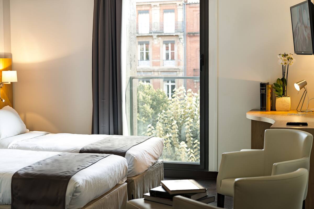 A hotel room with two beds, a desk with a lamp, a chair, and a view of a building and trees outside the window.