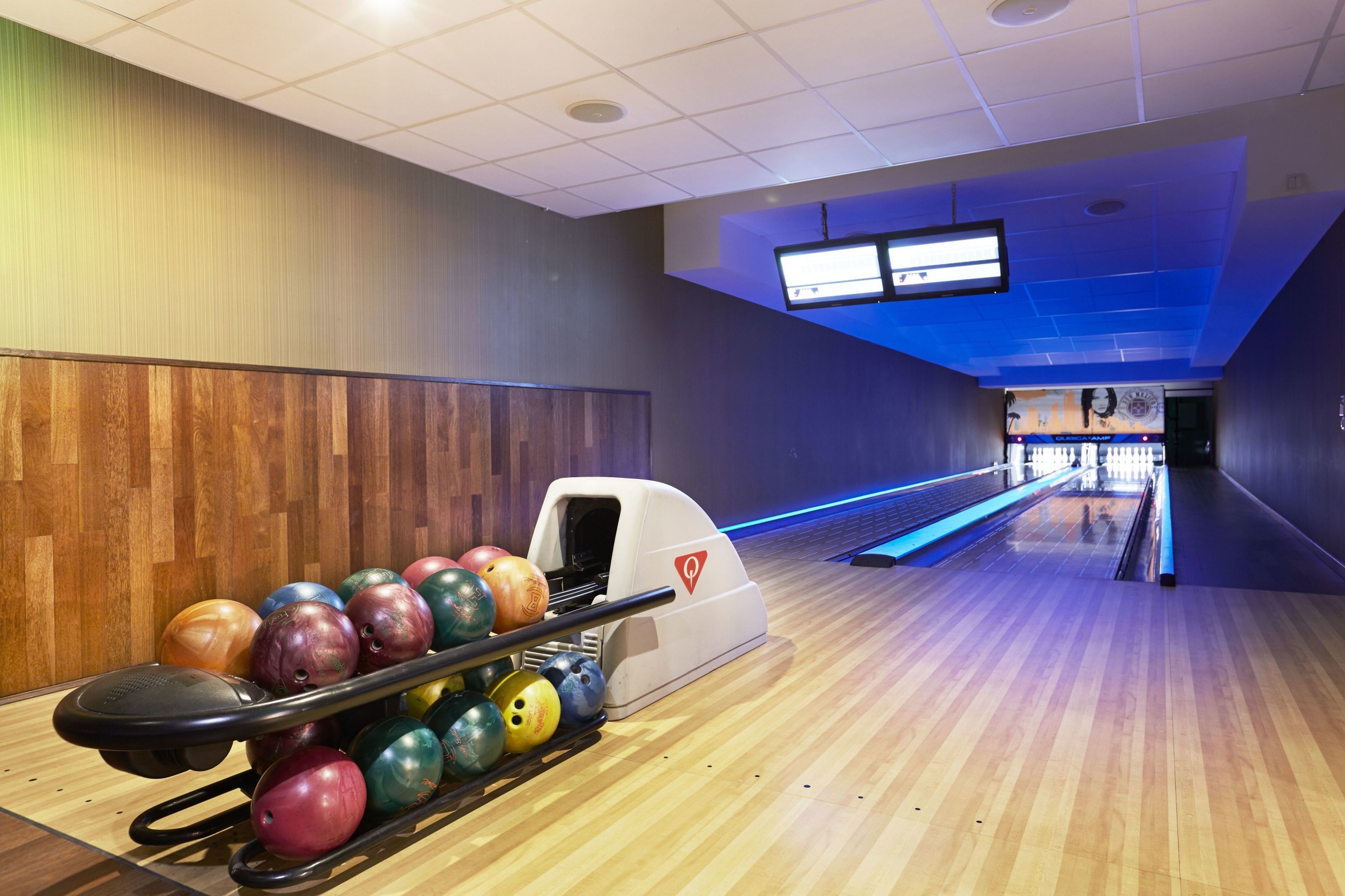 A bowling alley with a wooden floor, a bowling ball return, and a TV screen on the wall.