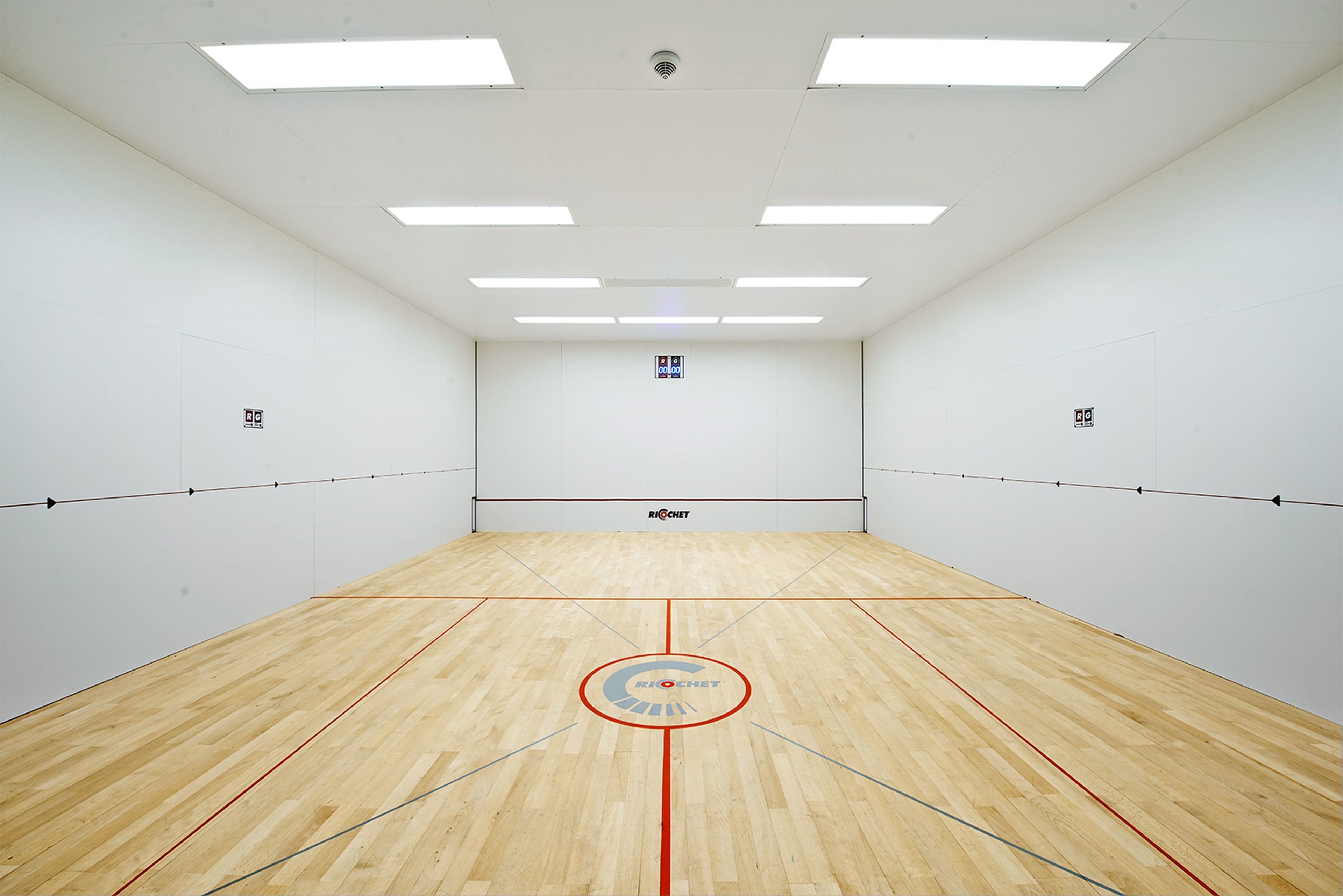 Una cancha de squash con piso de madera, paredes blancas e iluminación en el techo.