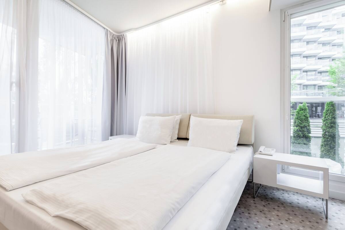 Una habitación de hotel con una cama grande, una mesita de noche y una ventana con vistas a edificios y árboles.