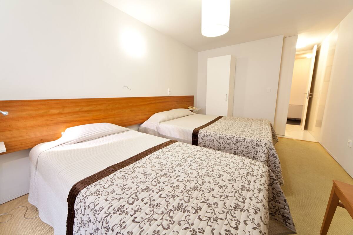 A hotel room with two beds, a wooden headboard, a bedside table with a lamp, and a door leading to another room.