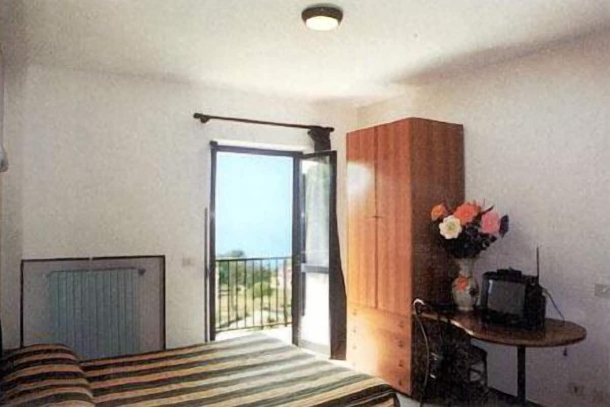 Una habitación con cama, un armario de madera, una mesita con un florero y vistas a través de un ventanal.