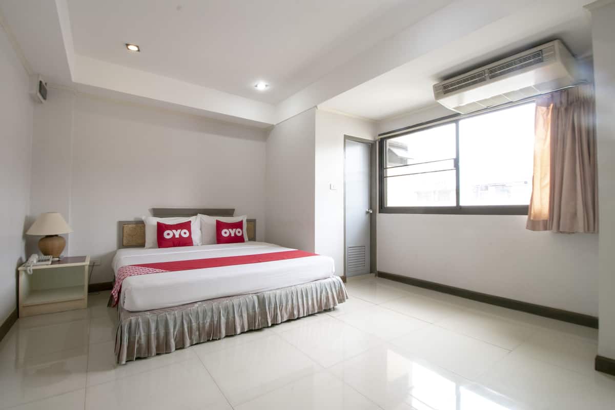 A hotel room with a large bed, a bedside table with a lamp, a window with curtains, and an air conditioning unit.
