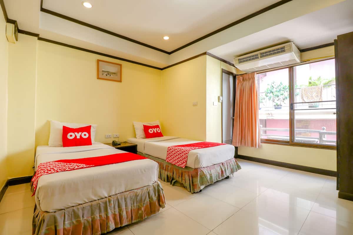 A hotel room with two beds, each with a red and white bedspread and pillows, a window with a view of the outside, and an air conditioning unit on the wall.
