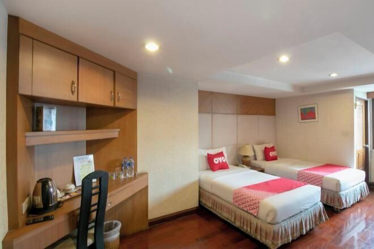 A hotel room with two beds, a desk with a chair, a small kitchenette, and a window.