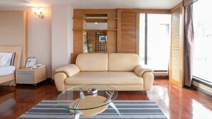 A modern hotel room with a beige sofa, a glass coffee table, a wooden cabinet, and a large window with blinds. - Hotel Win Long (Bangkok)