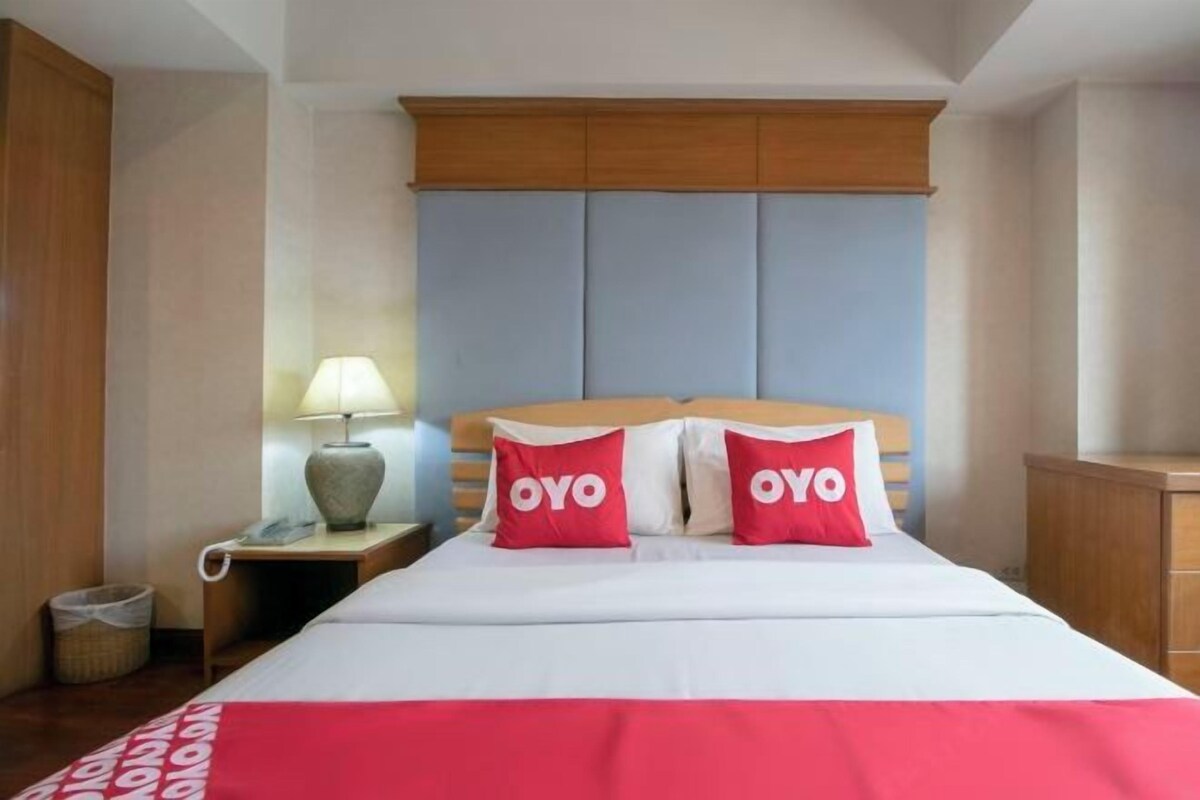 A hotel room with a bed, two pillows with 'OYO' branding, a nightstand with a lamp, and a wooden wardrobe.