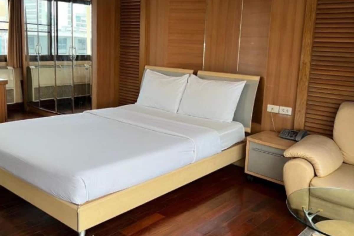 A modern hotel room with a wooden paneled wall, a large bed, a glass table, and a chair.