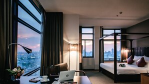 Habitación de hotel con escritorio, laptop y periódico, cama con almohadas y vista del paisaje urbano a través de amplias ventanas.