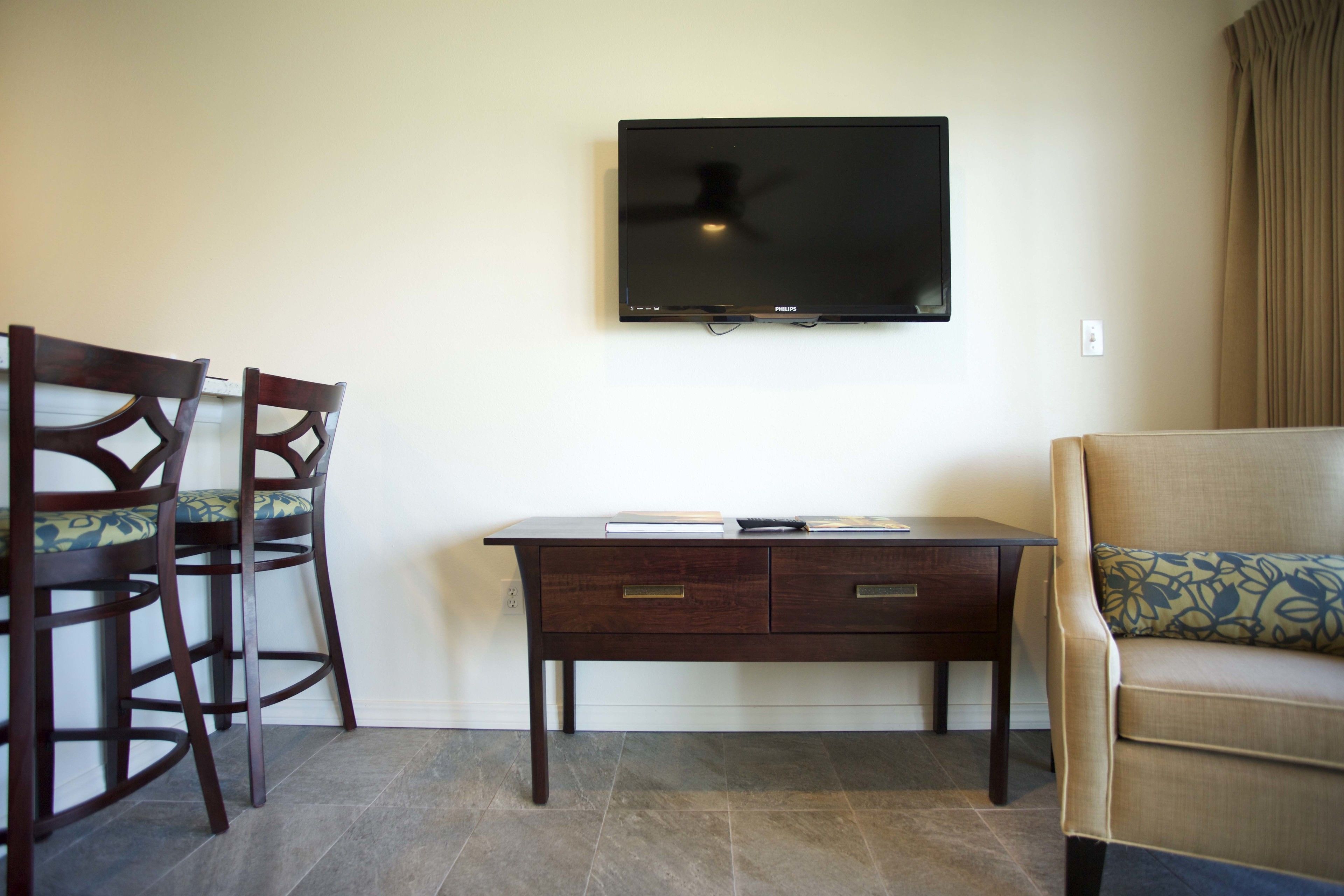 Habitación con televisión de pantalla plana en la pared, escritorio con dos cajones, dos sillas y sofá.