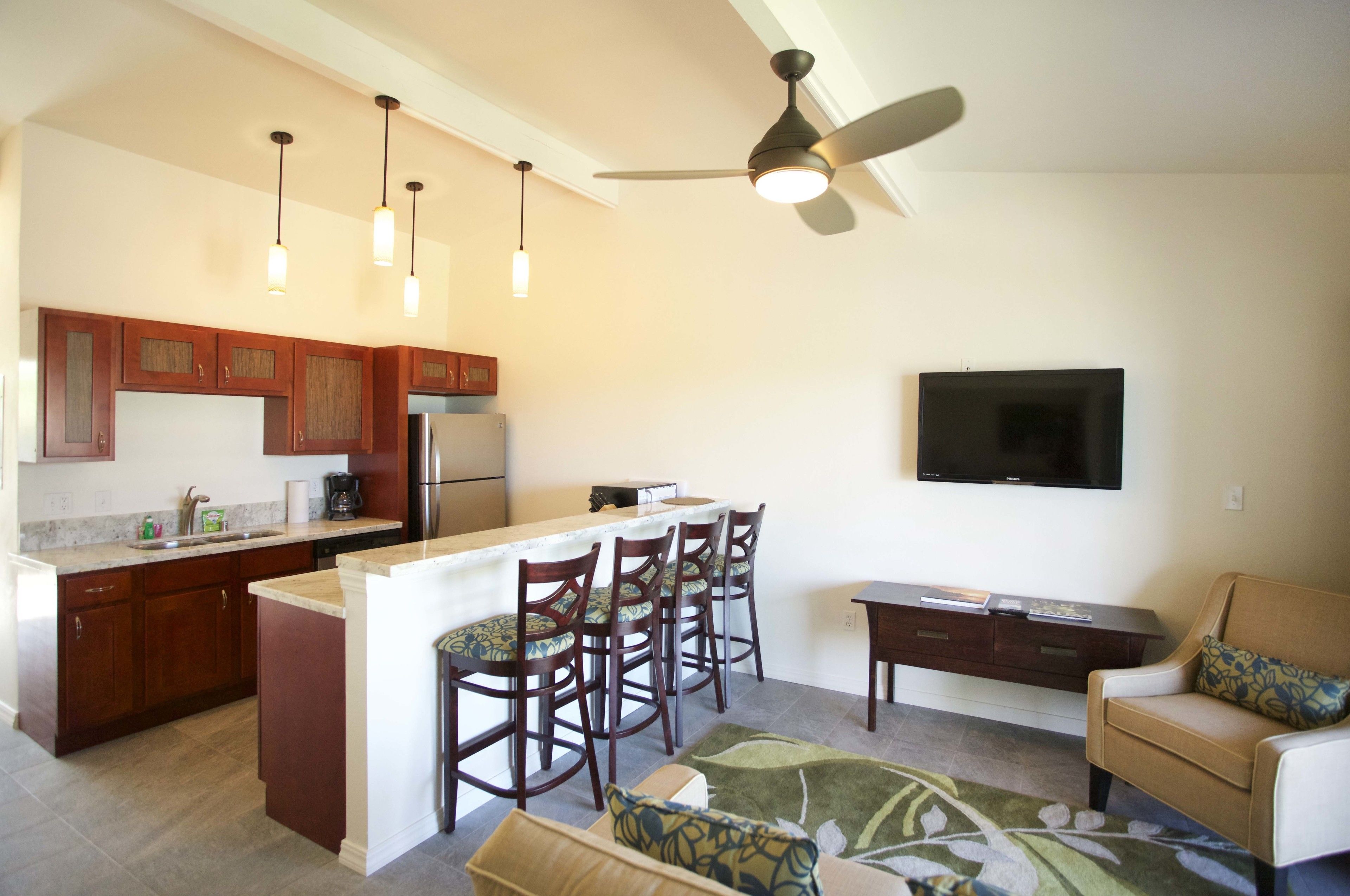 A modern kitchen with a central island, bar stools, a dining table, and a flat-screen TV mounted on the wall.