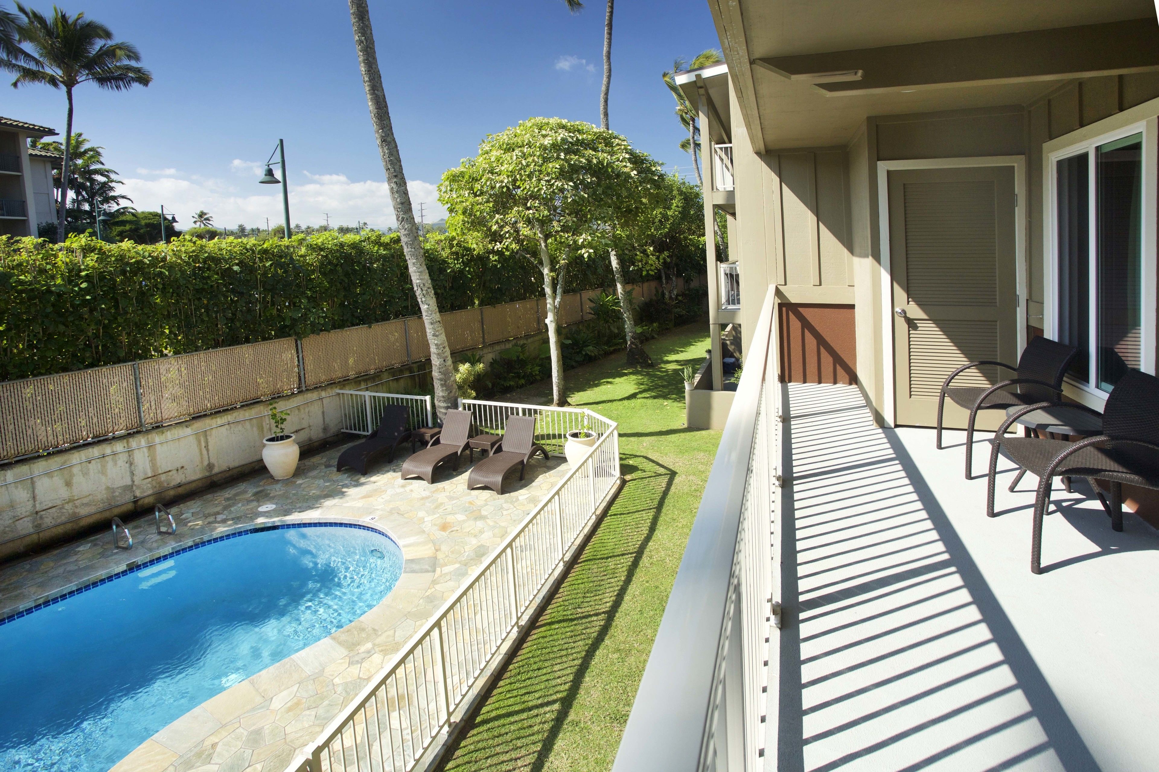A swimming pool with a stone edge, surrounded by a white fence, and a deck with chairs.