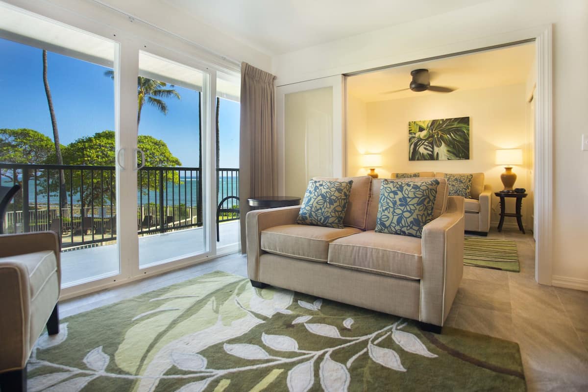 A living room with a sofa, a green patterned rug, a painting, and a view of the ocean through large sliding doors.