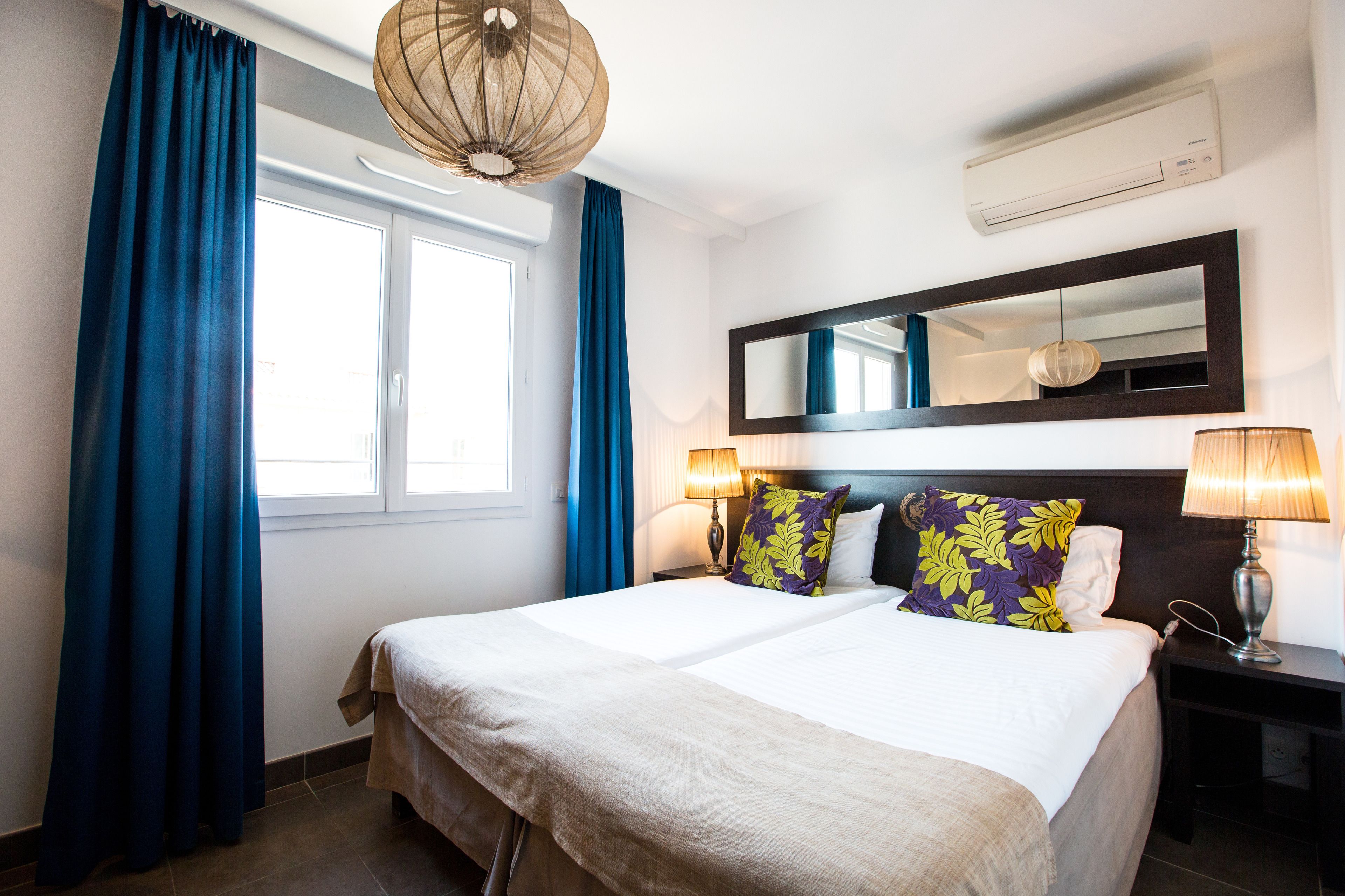 A hotel room with a large bed, two bedside lamps, a mirror above the bed, and a window with blue curtains.
