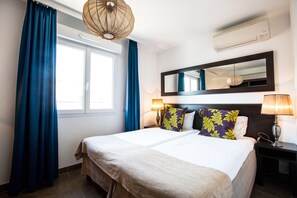 Une chambre d’hôtel avec un grand lit, deux lampes de chevet, un miroir au-dessus du lit et une fenêtre avec des rideaux bleus.