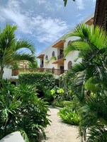 Un cortile con un viale in pietra, circondato da una vegetazione rigogliosa e un edificio bianco con balconi.