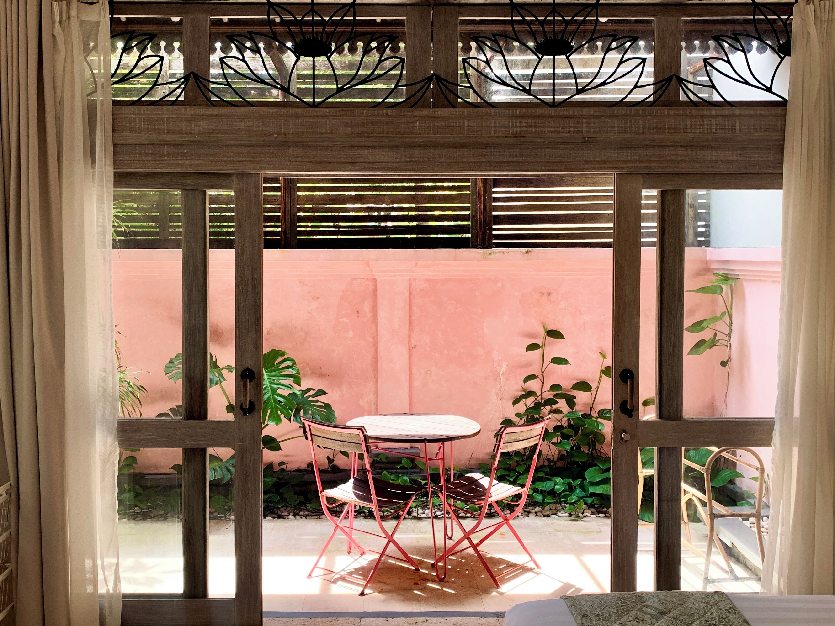 Porta aberta dando para um pátio com mesa e cadeiras, parede rosa e vegetação.