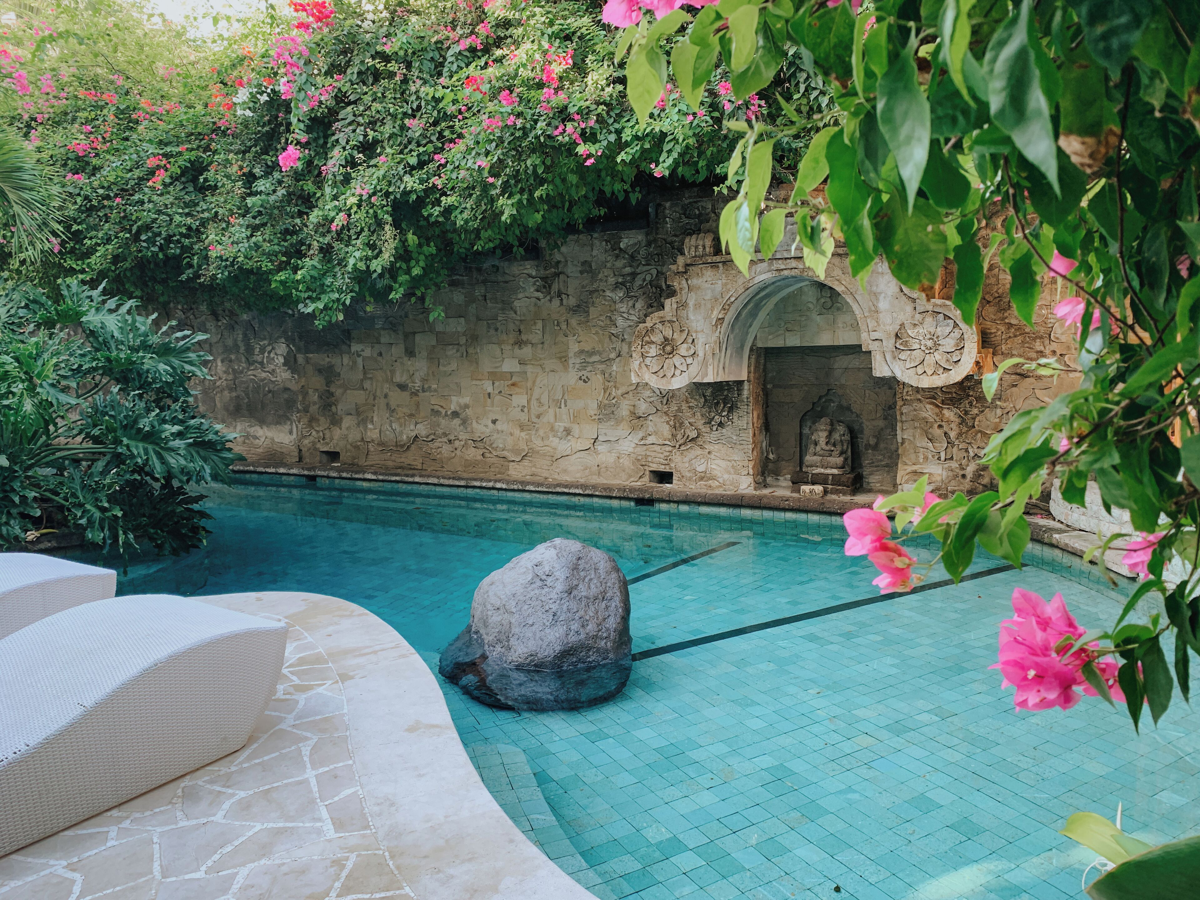 A serene pool area with a stone wall, a Buddha statue, and lush greenery.