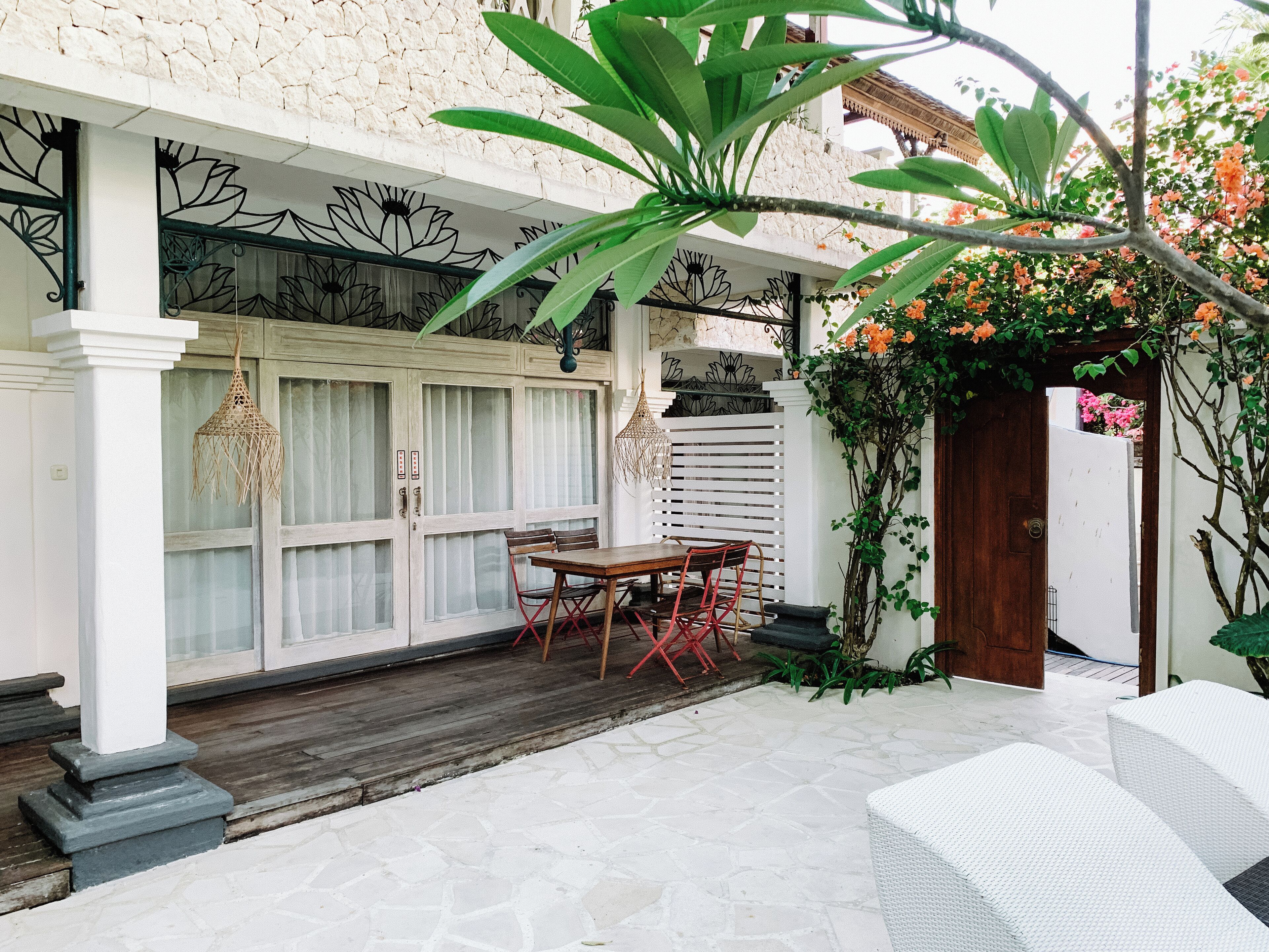 Un patio techado con una mesa y sillas de madera, una puerta blanca y piso de piedra.