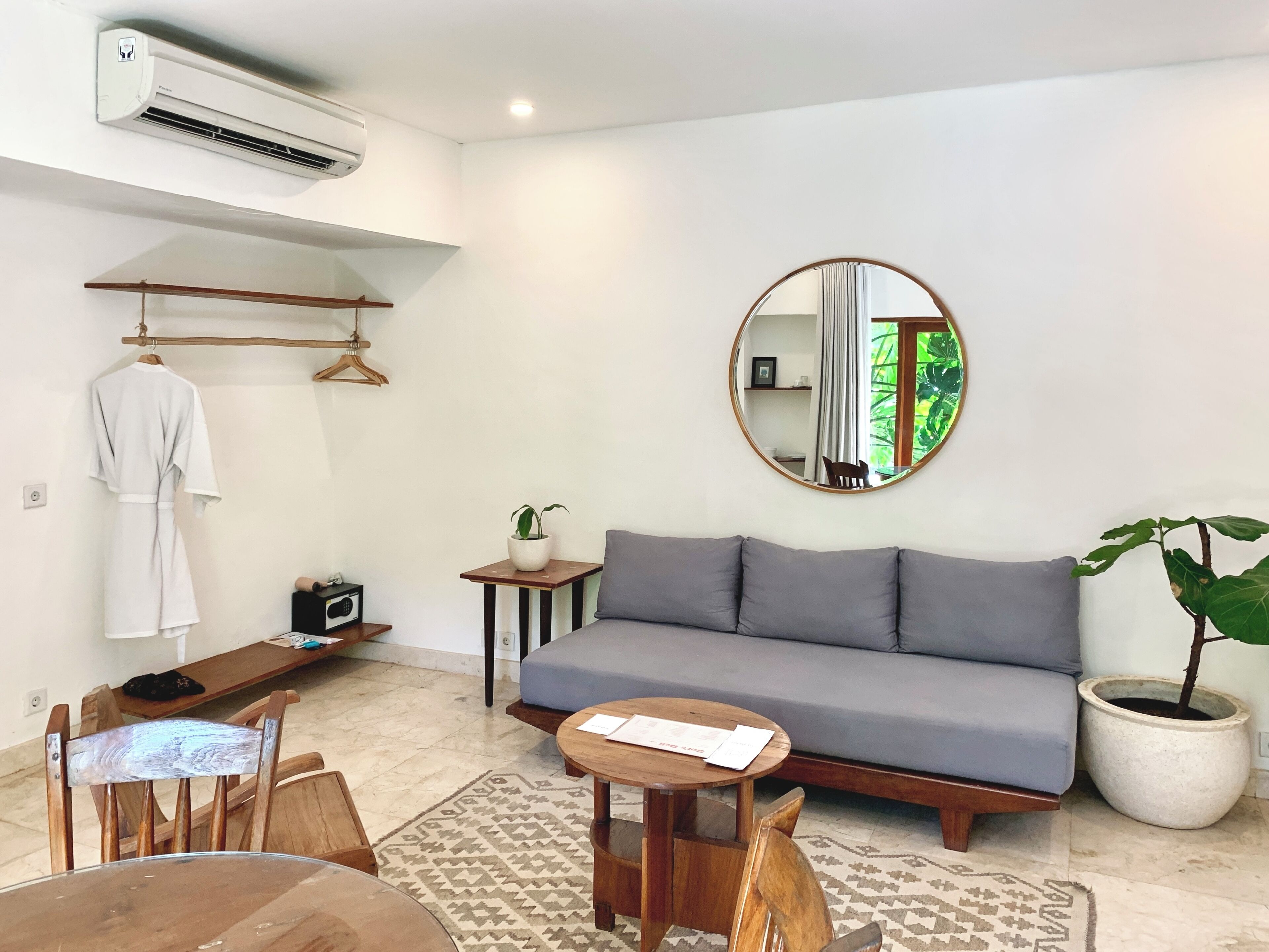 A living room with a grey sofa, a round mirror, a wooden table, a hanging robe, and a potted plant.