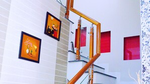 Una escalera con barandillas de vidrio y pasamanos de madera, flanqueada por paredes blancas con ventanas de tonos rojizos.