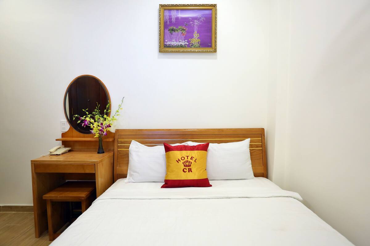 A hotel room with a wooden headboard, a bed with white bedding, a red and yellow pillow with "HOTEL CR" text, a wooden nightstand with a phone, a vase with flowers, and a framed picture on the wall.