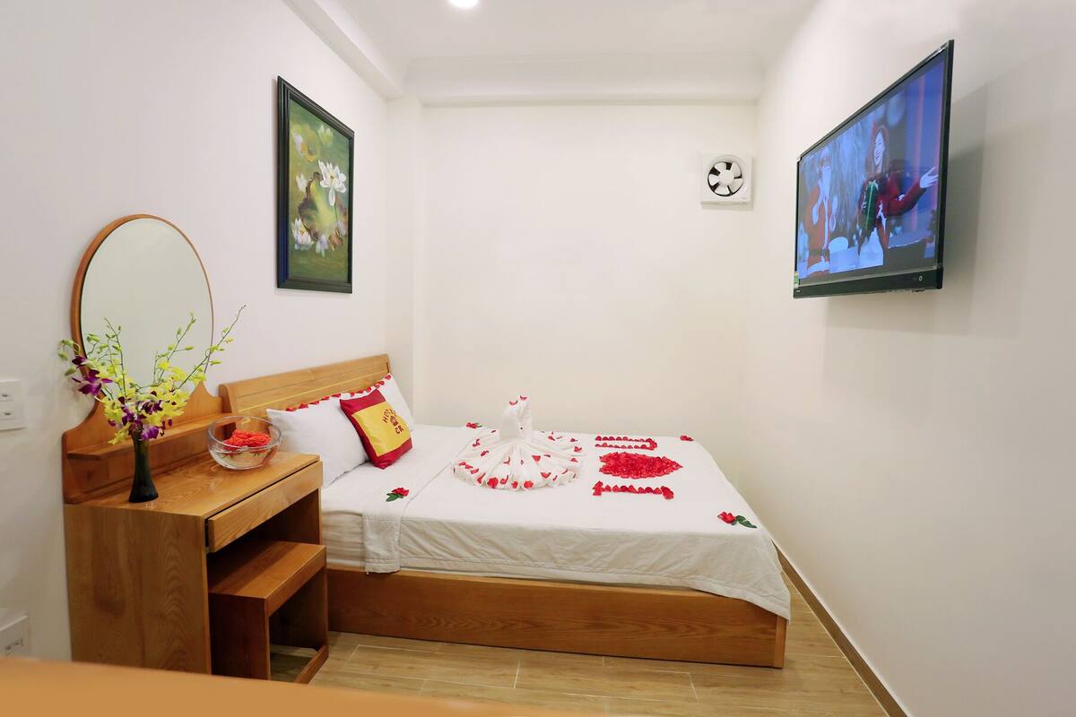 A neatly arranged bedroom with a wooden bed, a bedside table with a vase of flowers, a wall-mounted TV, and a framed painting.
