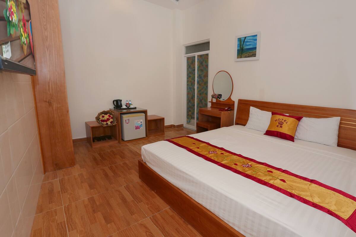 A hotel room with a large bed, a wooden nightstand, a small desk with a mirror, and a door leading to a balcony.