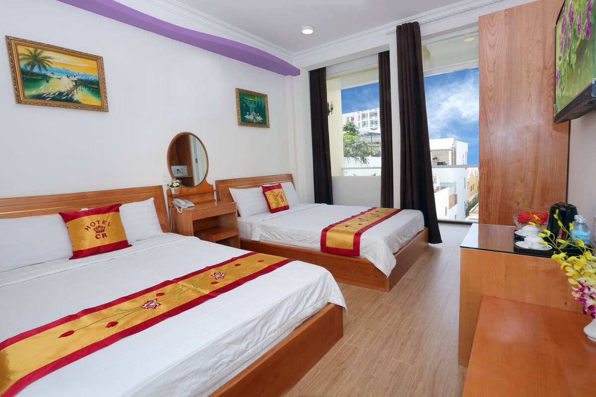 A hotel room with two beds, a wooden desk, a mirror, and a window with curtains.