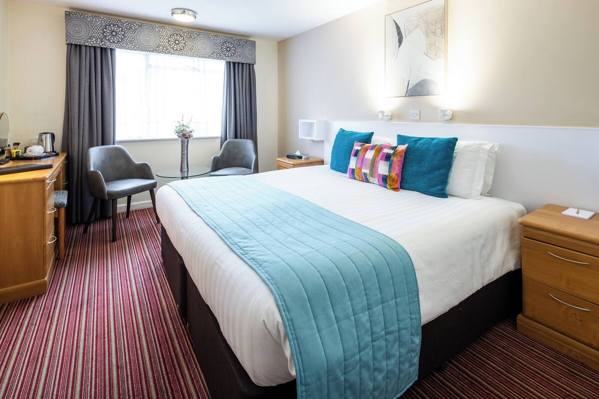 Hotelkamer met een groot bed, een bureau met stoel, een kleine tafel met vaas, en een raam met gordijnen.
