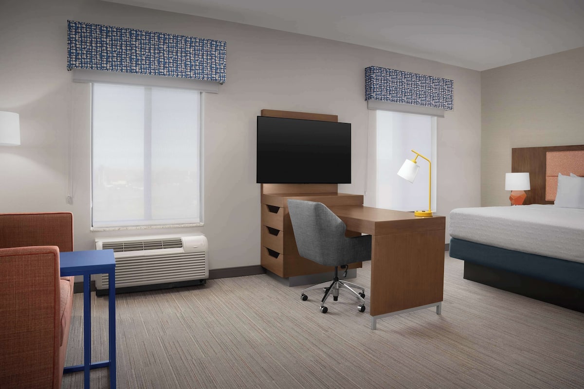 A hotel room with a bed, a desk with a chair, a television, and a window with blinds.