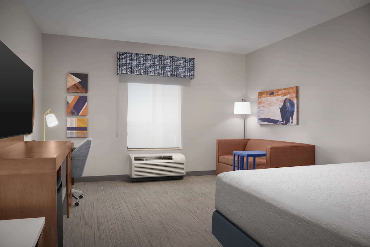 A hotel room with a bed, a desk, a chair, a lamp, and artwork on the walls.