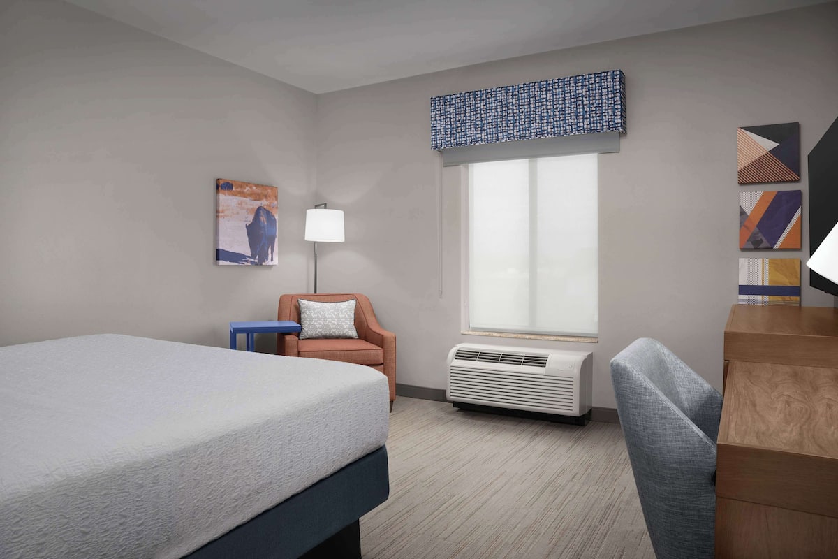 A hotel room with a bed, a chair, a desk, a window with a view, and a wall-mounted air conditioner.