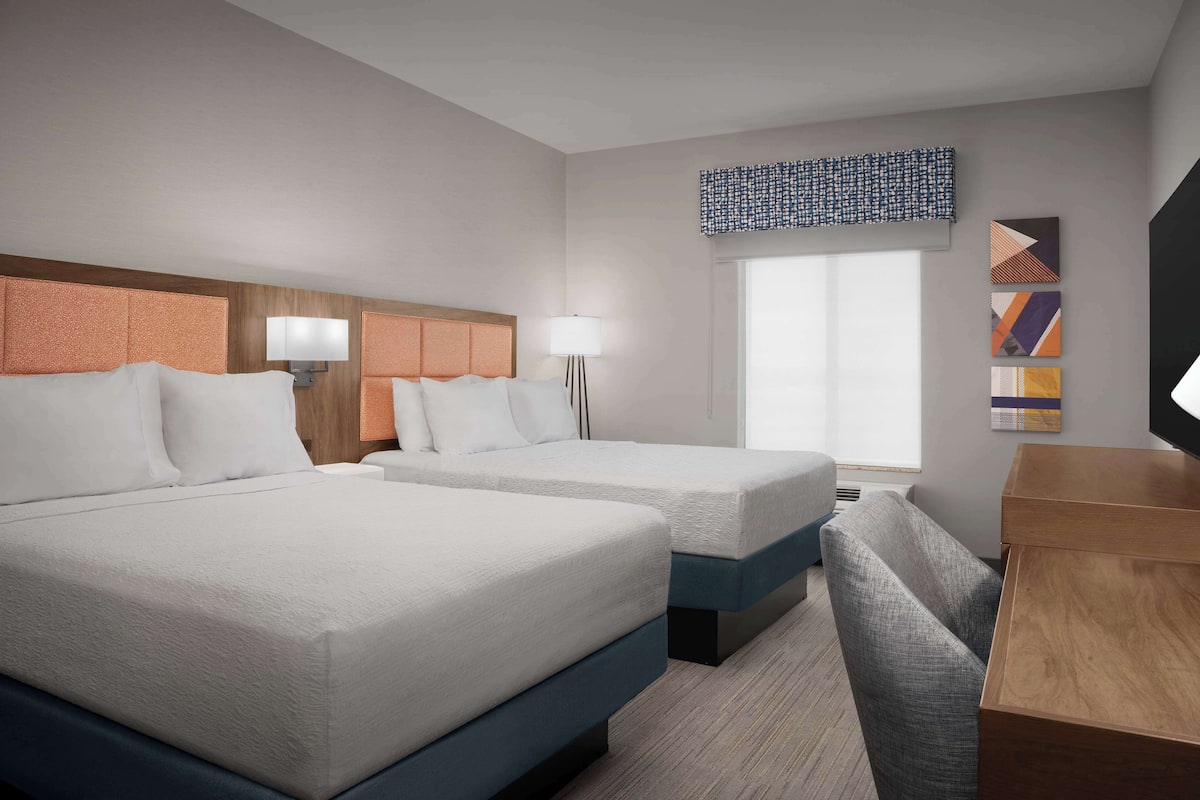 A hotel room with two beds, a desk, a chair, a lamp, and a window with a view.