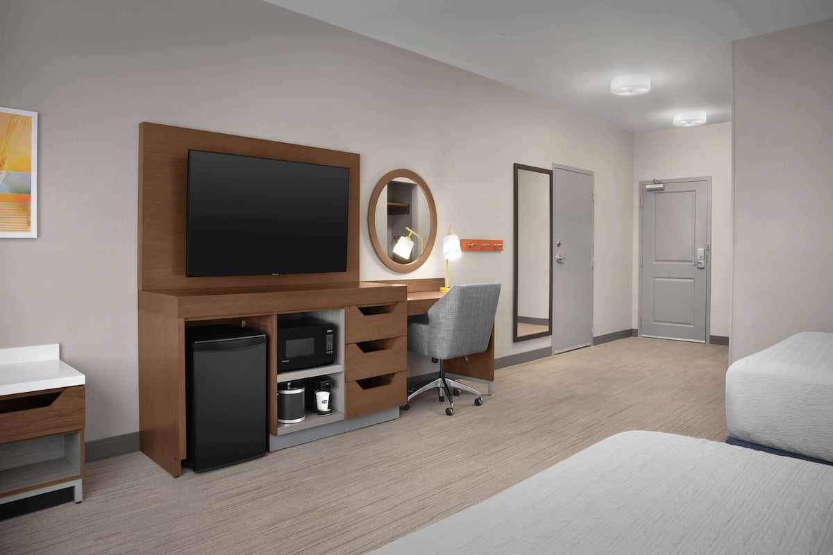 A hotel room with a flat-screen TV, a desk with a chair, a small refrigerator, a bedside table, and a door.