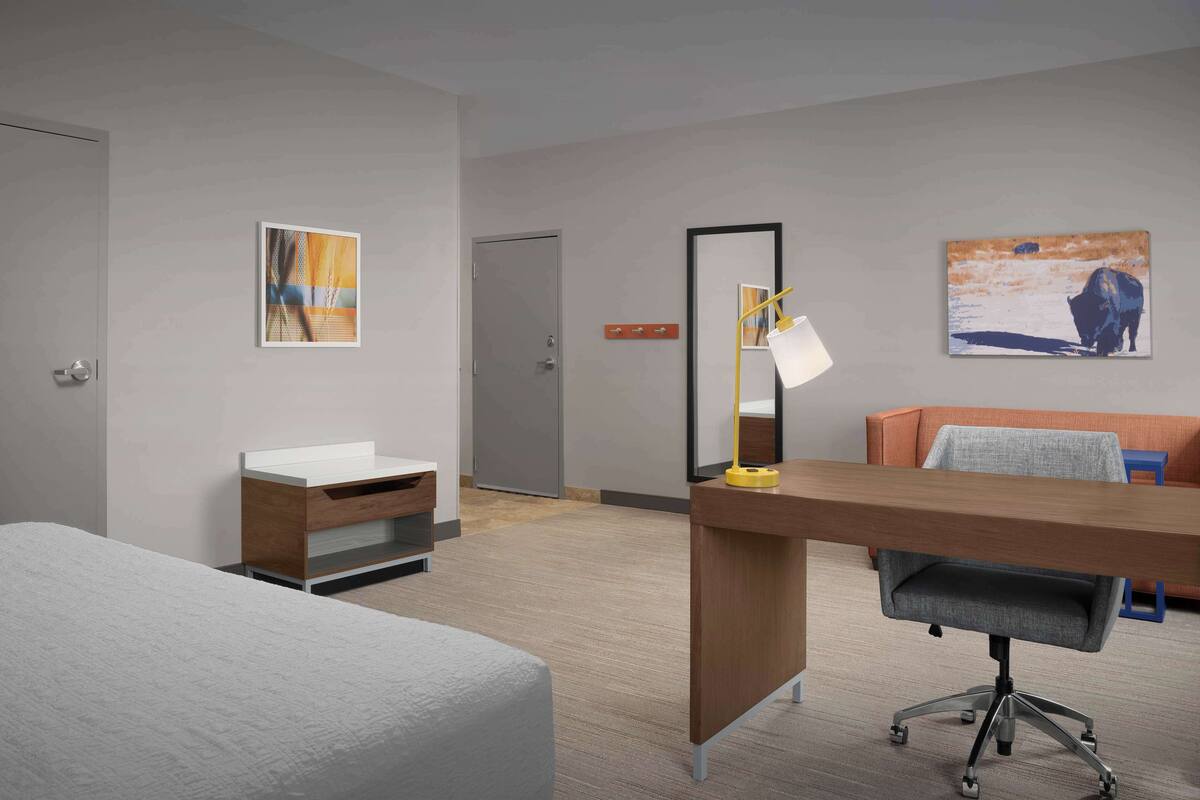 A hotel room with a bed, a desk with a chair, a mirror, and two paintings on the wall.