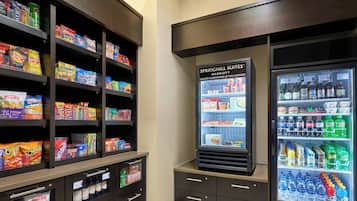 A hotel pantry with shelves stocked with snacks and beverages, a refrigerated display case, and a freezer.