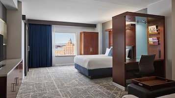 Habitación de hotel con una cama grande, un escritorio con silla, televisión y vista a la ciudad.