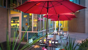An outdoor dining area with tables and chairs, red umbrellas, and a glass door entrance.