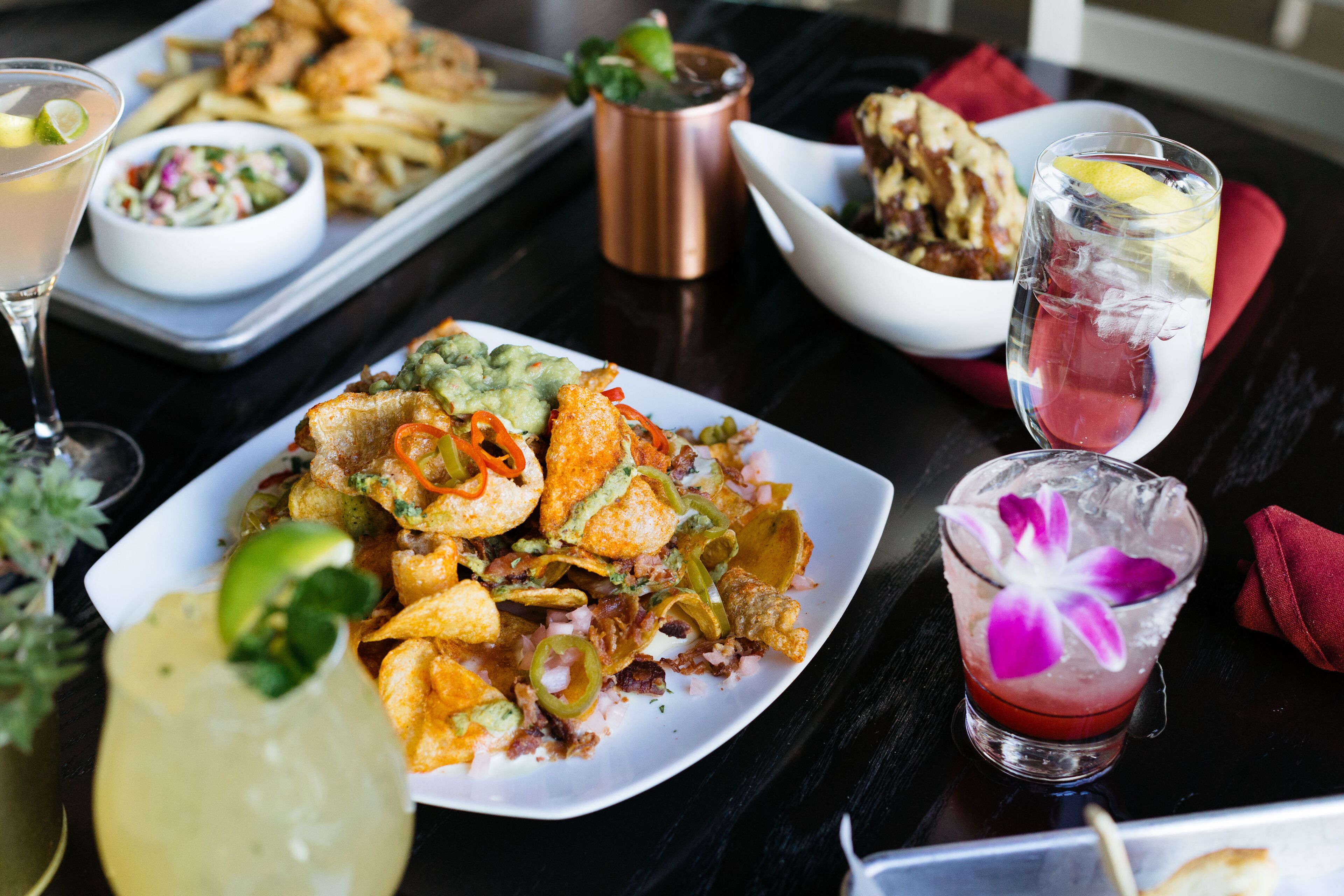 Uma mesa de jantar com diversos pratos, incluindo um prato de nachos, uma tigela de salada de repolho e uma taça de martini com uma flor.