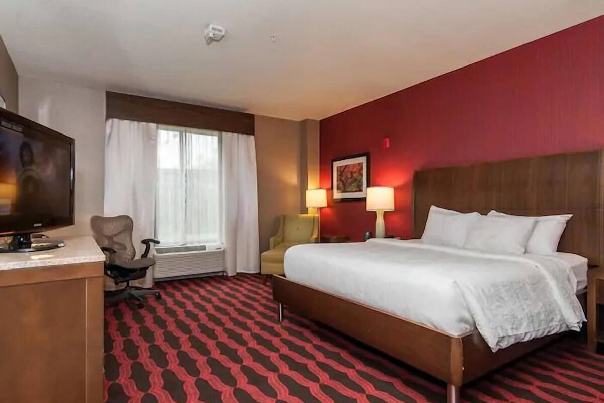 Une chambre d’hôtel comprenant un lit, un bureau avec une télévision, une chaise, une fenêtre avec des rideaux et deux lampes de chevet.