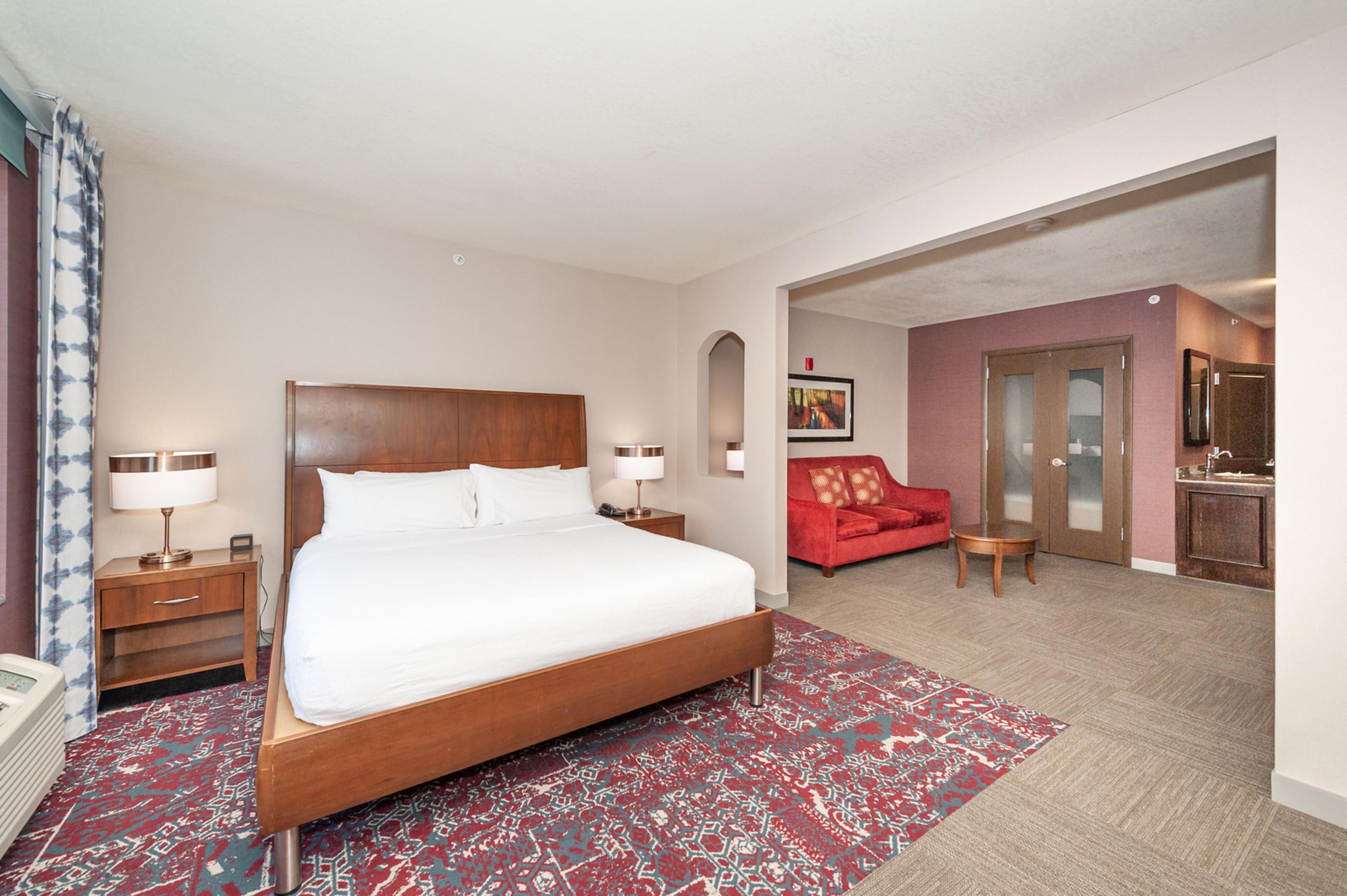 Una habitación de hotel con una cama grande, un sofá rojo, una mesita y un baño visible a través de un arco.