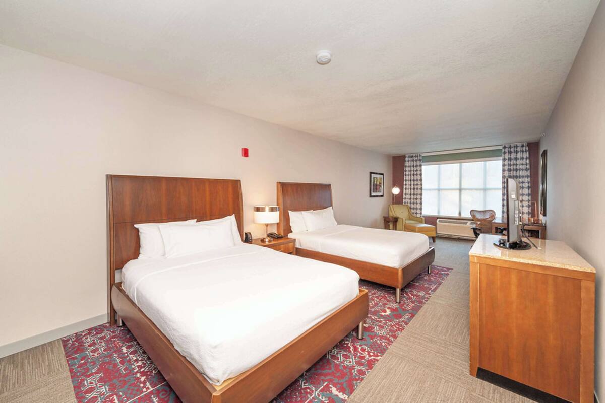 Une chambre d’hôtel avec deux lits, un bureau, une chaise, une fenêtre avec des rideaux et un tapis rouge à motifs.