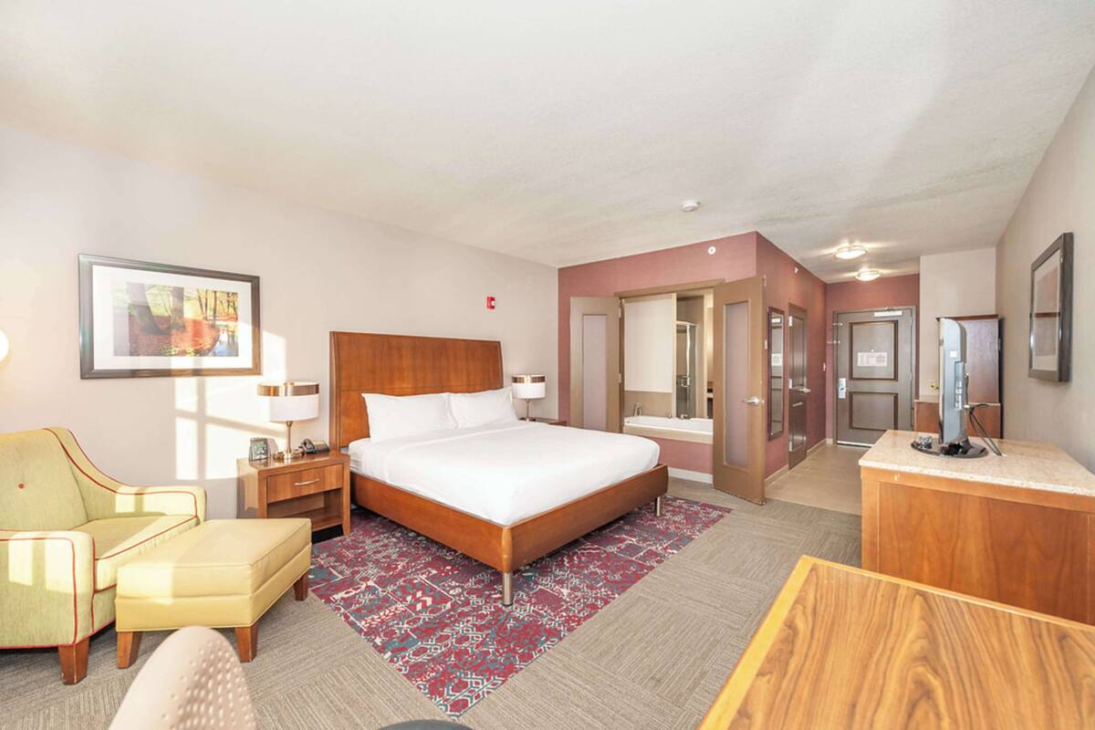 Une chambre d’hôtel avec un grand lit, un bureau, une chaise, une table de chevet, une lampe, un miroir et une porte de salle de bain.