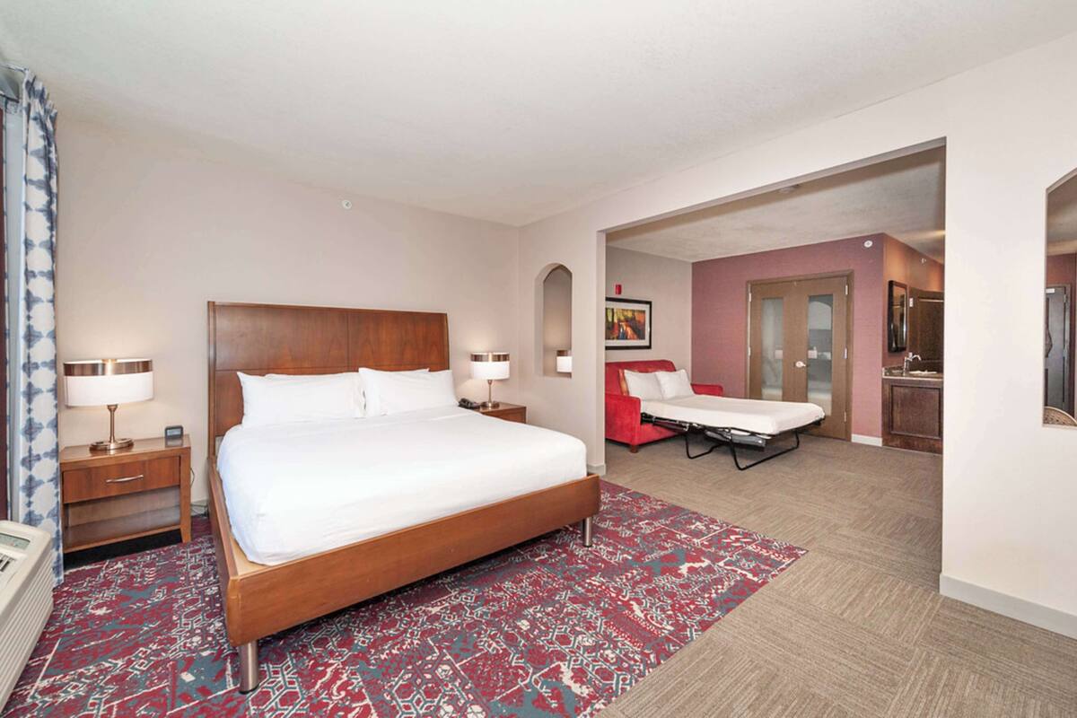 Une chambre d’hôtel avec un grand lit, des tables de chevet, un tapis rouge à motifs et un canapé rouge.
