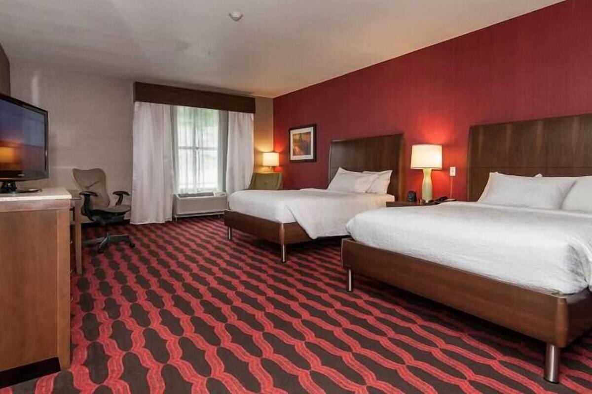 Une chambre d’hôtel avec deux lits, un bureau avec une télévision, une chaise et une fenêtre avec des rideaux.