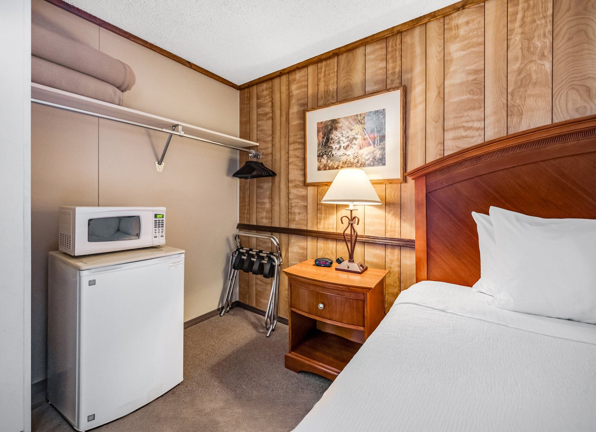 A hotel room with a bed, a bedside table with a lamp, a microwave, a refrigerator, and a coat rack.