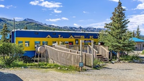 A blue and yellow building with 'ALASKA' written on it, a wooden staircase leading to a door, and a mountainous backdrop. - Denali Park Hotel (Healy)