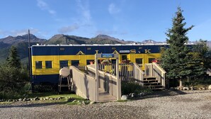 Estação de trem azul e amarela com uma escadaria de madeira que leva a uma plataforma, cercada por montanhas e vegetação.