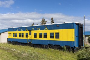 Un vagón de tren estacionado sobre rieles, con la palabra 'ALASKA' en su lateral, con césped y árboles de fondo.